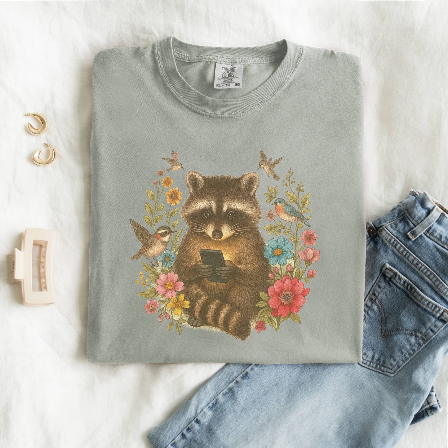 T-shirt design showing raccoon holding phone surrounded by colorful wildflowers and birds