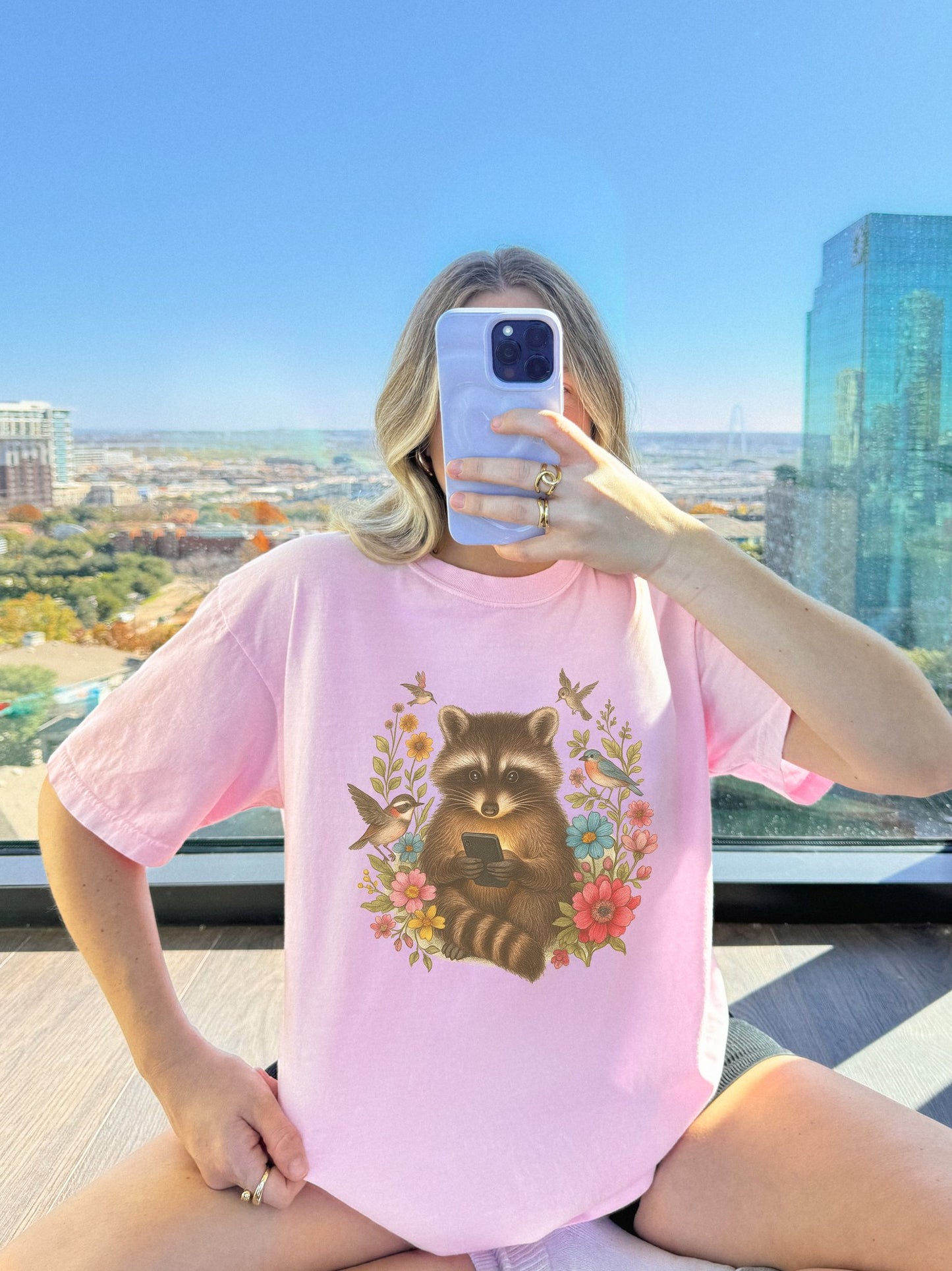 T-shirt design showing raccoon holding phone surrounded by colorful wildflowers and birds
