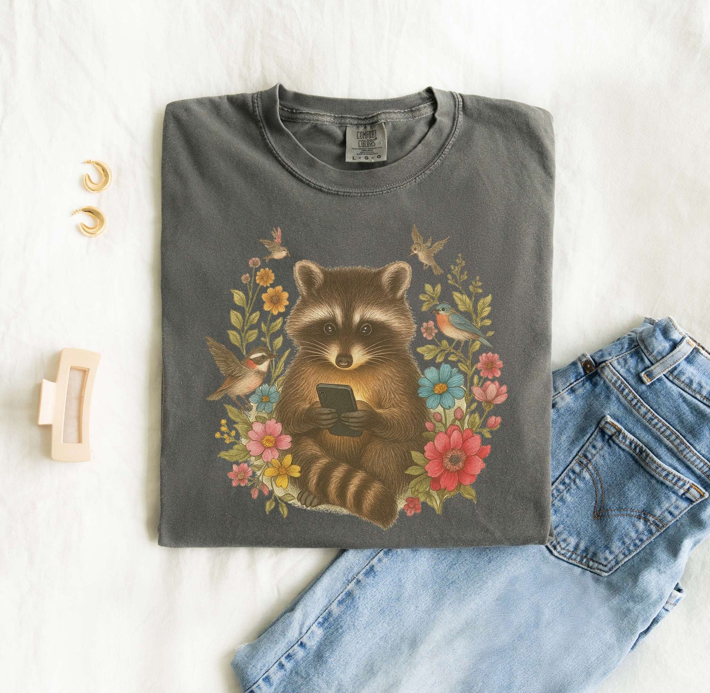 T-shirt design showing raccoon holding phone surrounded by colorful wildflowers and birds