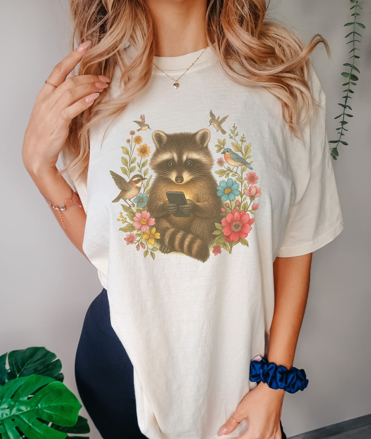 T-shirt design showing raccoon holding phone surrounded by colorful wildflowers and birds