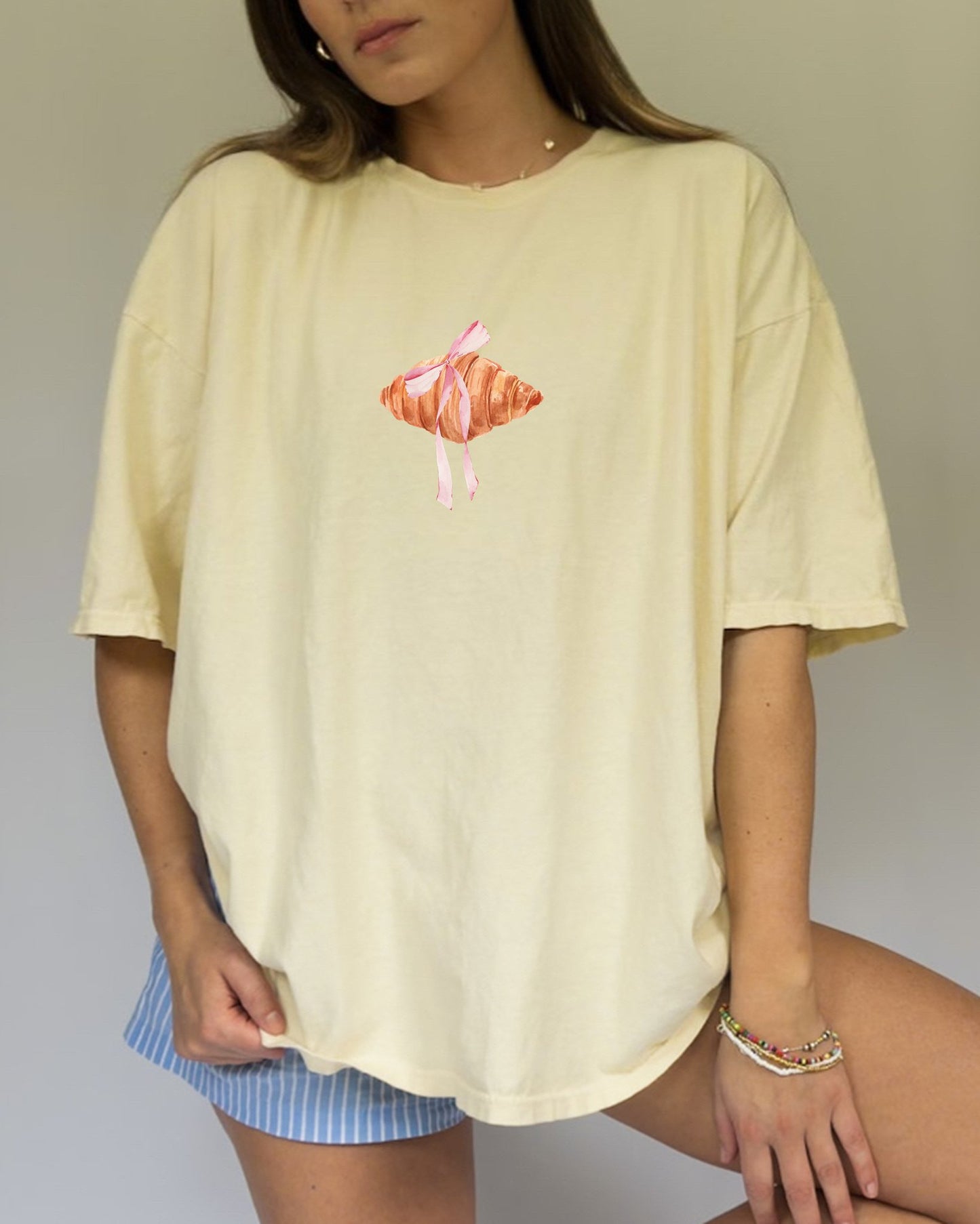 T-shirt with watercolor croissant design featuring golden brown pastry with pink ribbon bow