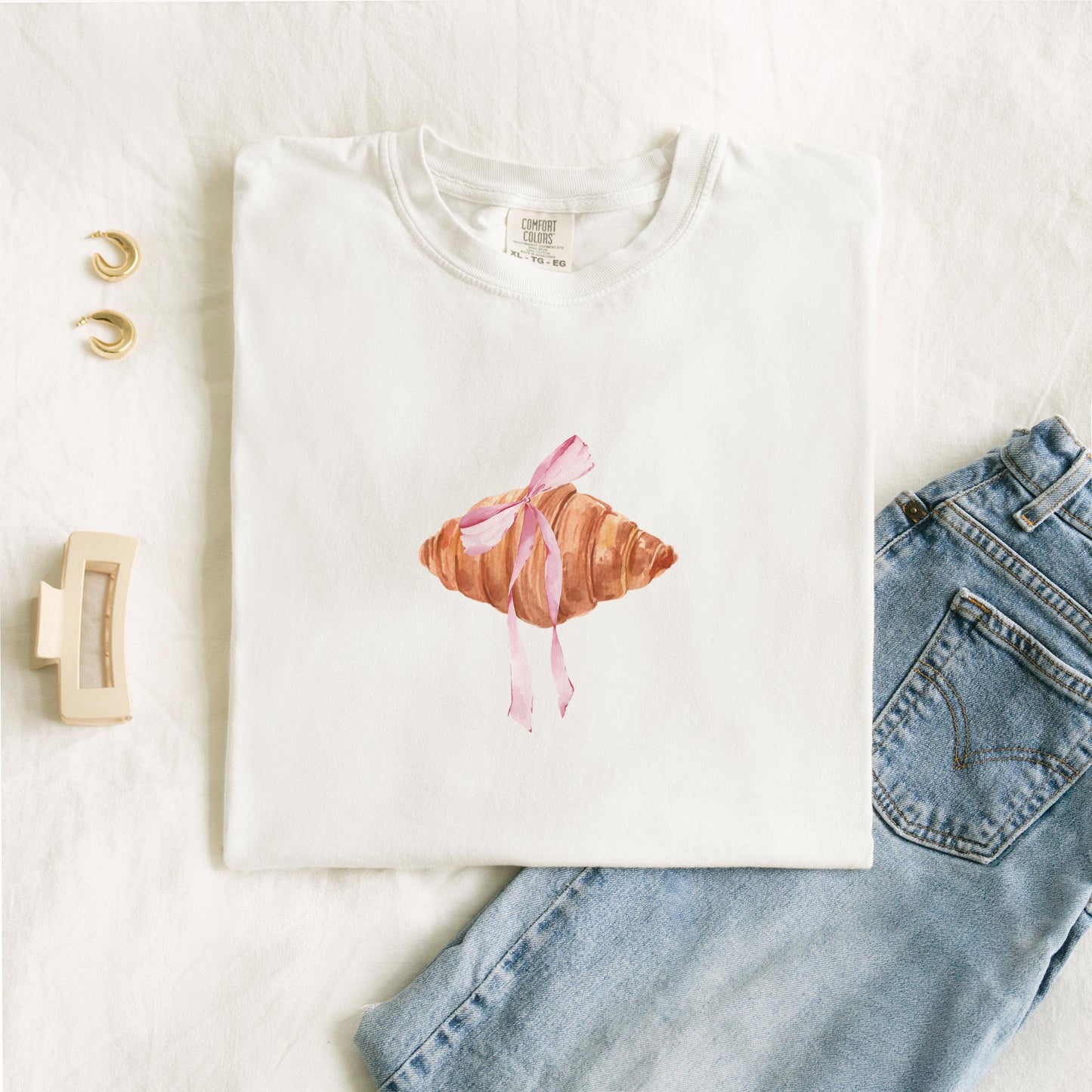T-shirt with watercolor croissant design featuring golden brown pastry with pink ribbon bow