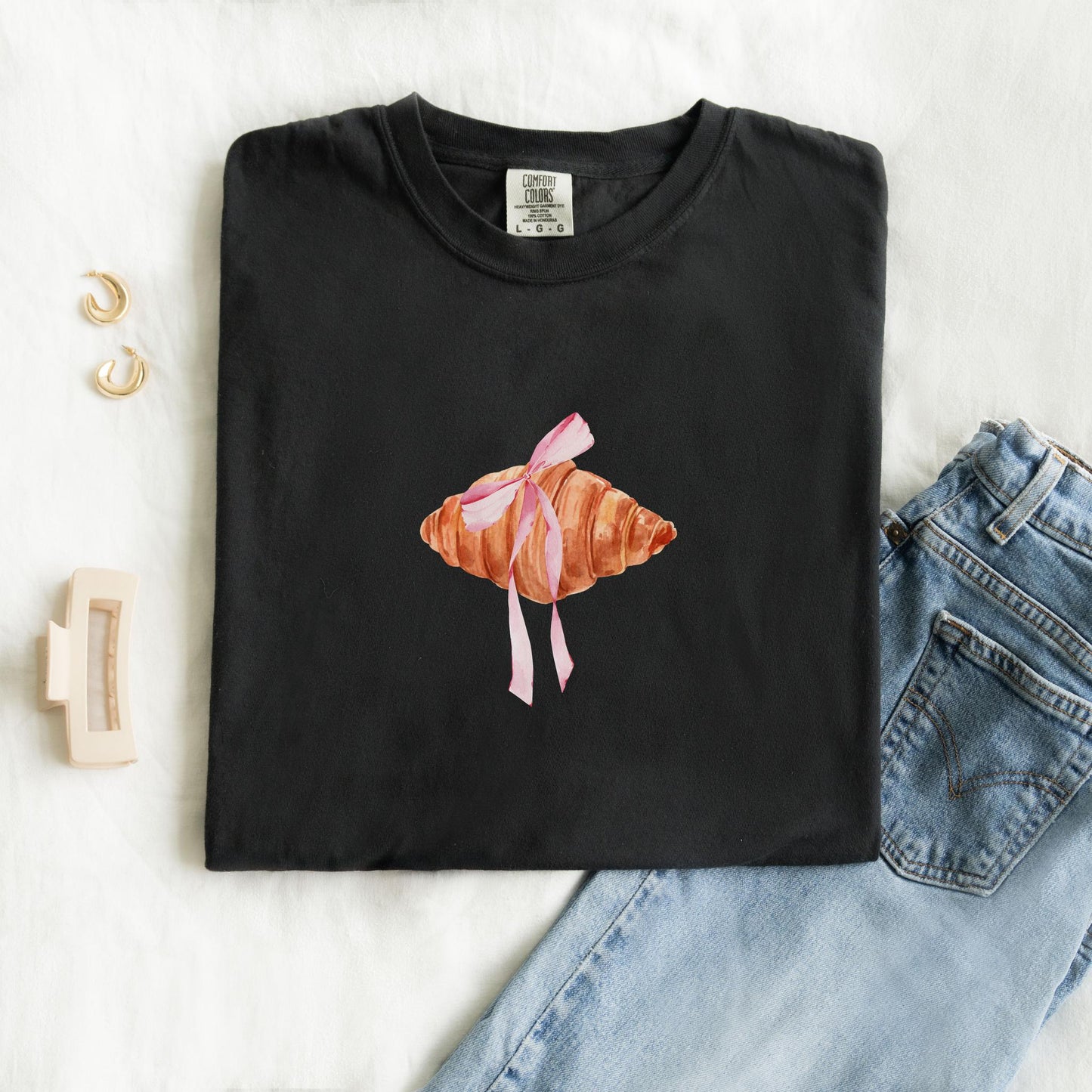 T-shirt with watercolor croissant design featuring golden brown pastry with pink ribbon bow