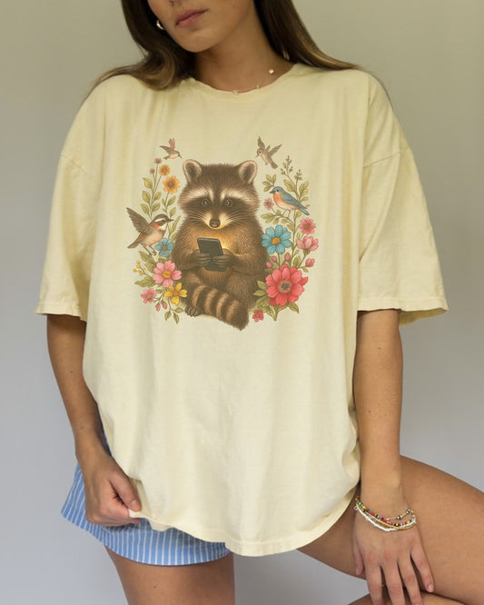 T-shirt design showing raccoon holding phone surrounded by colorful wildflowers and birds