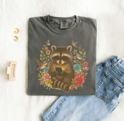 T-shirt design showing raccoon holding phone surrounded by colorful wildflowers and birds