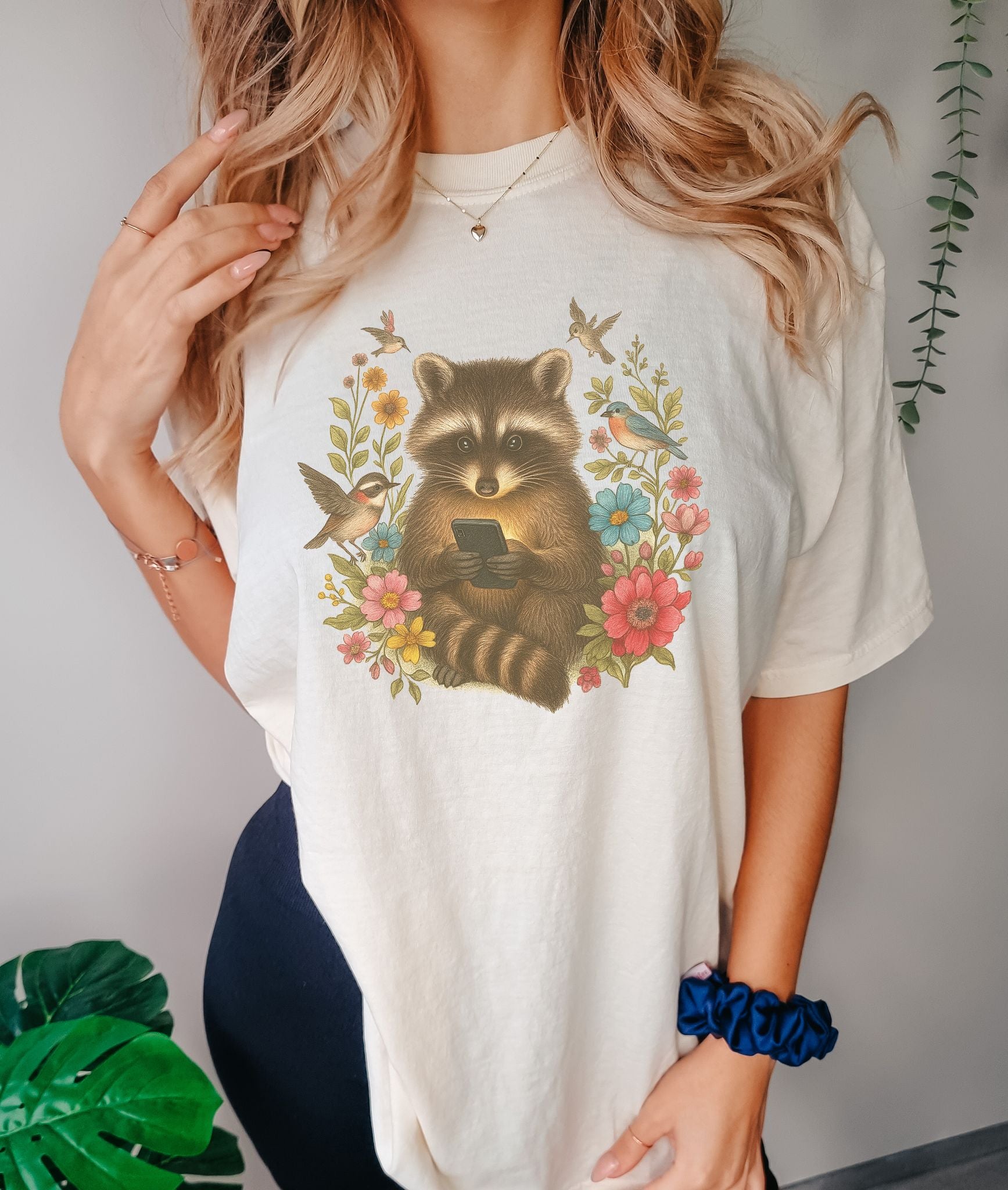 T-shirt design showing raccoon holding phone surrounded by colorful wildflowers and birds