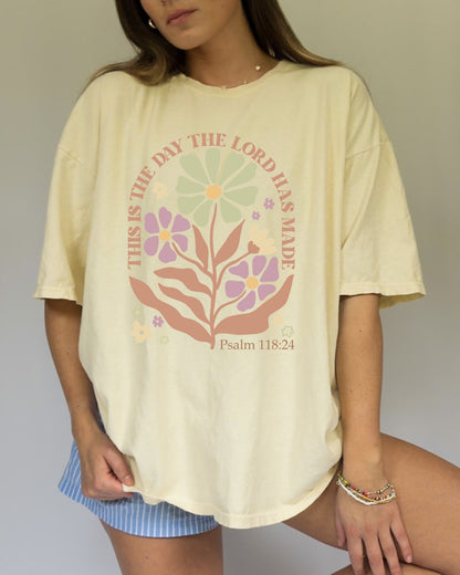 T-shirt with 'This is the Day' text, Psalm 118:24 verse, and floral design with protective hands in sage green, pink, and terracotta colors
