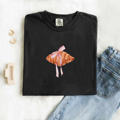 T-shirt with watercolor croissant design featuring golden brown pastry with pink ribbon bow