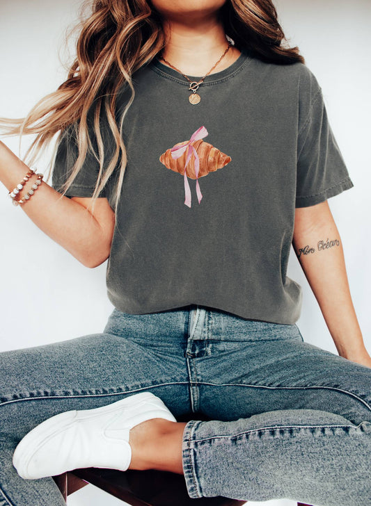 T-shirt with watercolor croissant design featuring golden brown pastry with pink ribbon bow